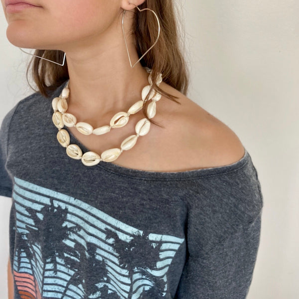 Model is wearing 2 separate white cowrie shell necklaces with different length settings so they look layered nicely with her island tshirt and heart silver hoop earrings.